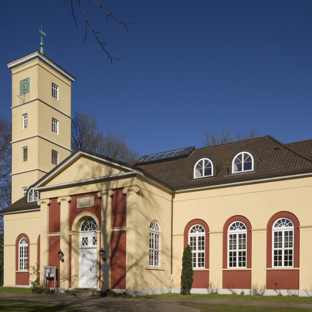 Das Bild zeigt ein historisches Gebäude mit einer beige-rot gestrichenen Fassade und einem schlanken Turm. Die Fassade hat große, bogenförmige Fenster und eine zentrale Eingangstür mit Stufen. Ein klarer blauer Himmel ist im Hintergrund zu sehen.